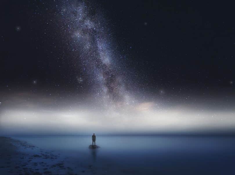 Silhueta de um homem em cima de uma pedra no meio do mar com um céu estrelado acima dele.