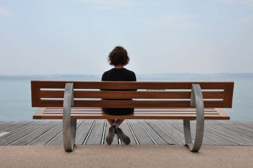 Pessoa sentada em um banco de madeira, em frente ao mar, em um dia nublado.