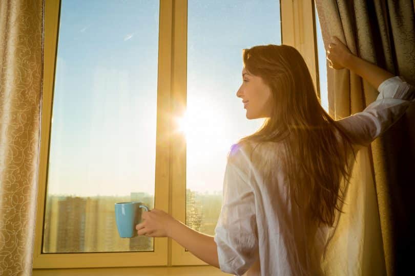 Mulher segurando uma xícara de café enquanto abre a cortina.
