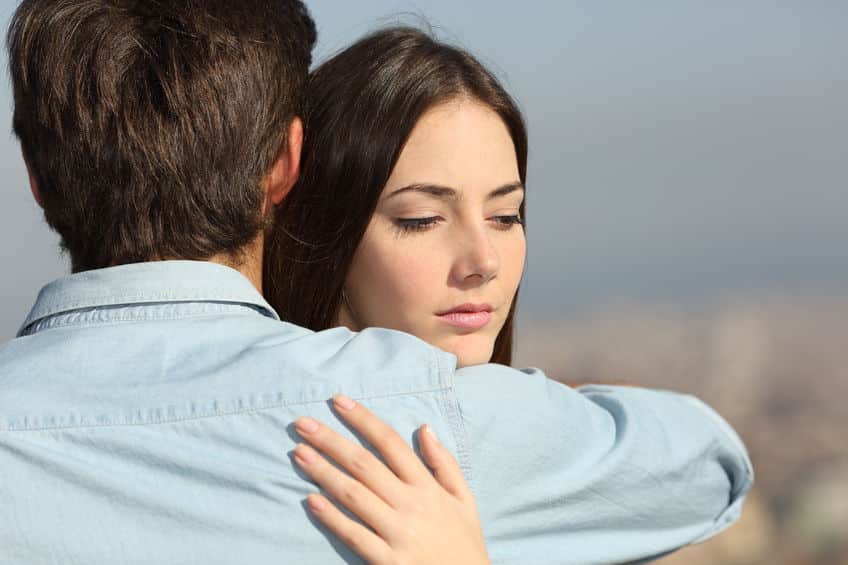 Mulher abraça homem enquanto olha para baixo, com expressão desconfiada.