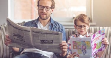 Homem e menino sentados em sofá com óculos lendo jornal e revista infantil