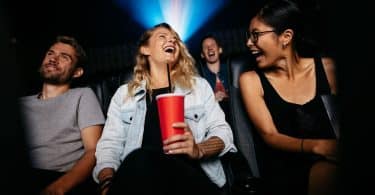 Duas mulheres e um homem sentados em uma sala de cinema sorrindo e demonstrando alegria.