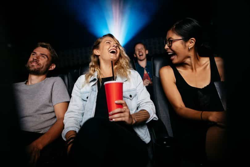 Duas mulheres e um homem sentados em uma sala de cinema sorrindo e demonstrando alegria.