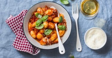 Prato de nhoque com molho, em cima de um pano quadriculado vermelho, com uma colher dentro e um garfo ao lado - acompanhado de um copo de bebida e um ramekin.