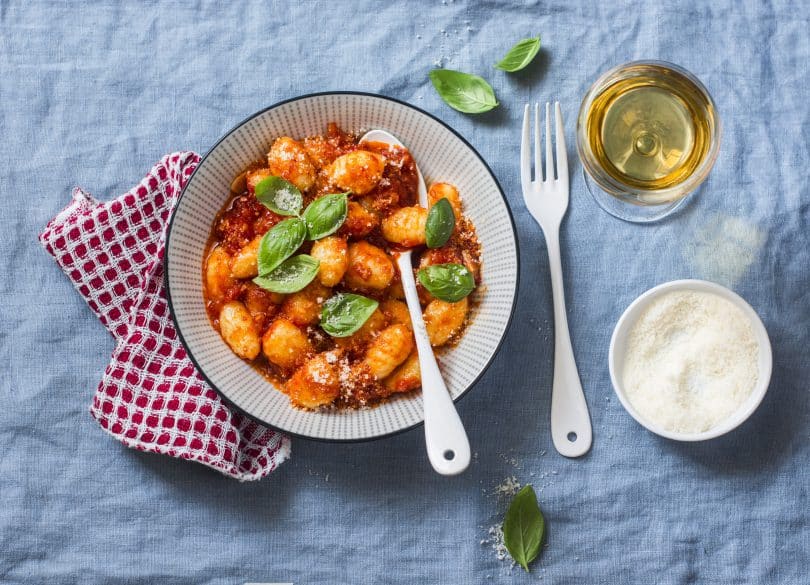 Prato de nhoque com molho, em cima de um pano quadriculado vermelho, com uma colher dentro e um garfo ao lado - acompanhado de um copo de bebida e um ramekin.