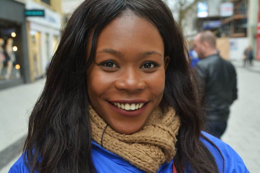 Imagem de uma linda mulher negra com seus cabelos pretos e longos. Ela está sorrindo e usa um cachecol marrom envolta do seu pescoço.
