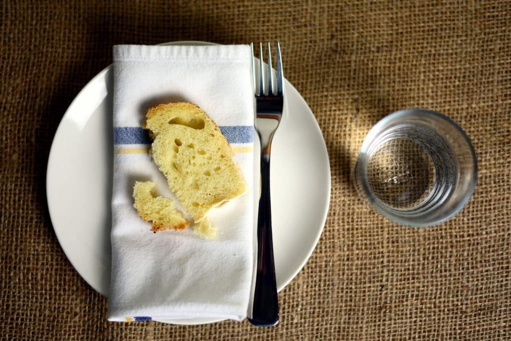 Imagem de uma mesa com uma toaha de juta. Sobre ela um prato branco com um guardanapo de boca sobre ele e um pedaço de pão e um garfo. Ao lado um copo com água.