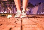 Imagem de um pé calçando um tênis. A pessoa está indo caminhar em uma noite bem gostosa e de muito calor. Ao lado do pés, uma flor branca.