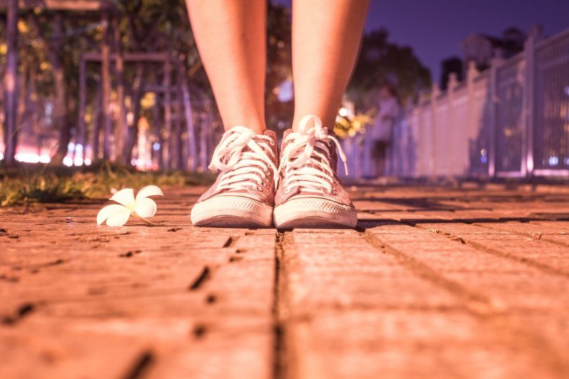 Imagem de um pé calçando um tênis. A pessoa está indo caminhar em uma noite bem gostosa e de muito calor. Ao lado do pés, uma flor branca.