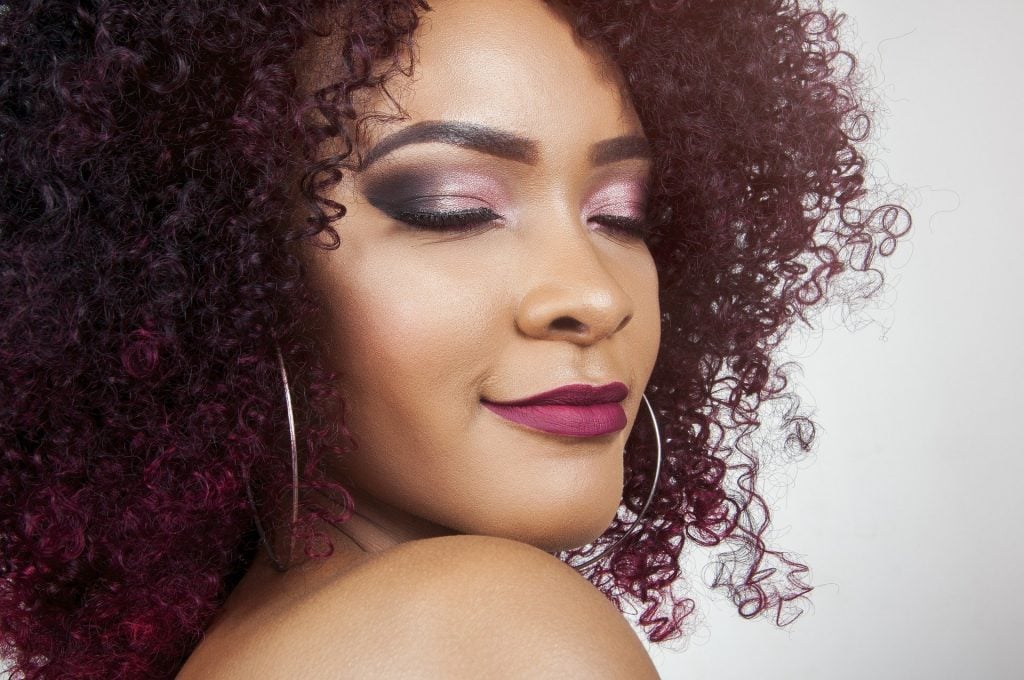 Imagem de uma bela mulher negra de cabelos cacheados na cor vermelho escuro. Ela está com os olhos e a boca pintados e usa um lindo e grande brinco de argolas.