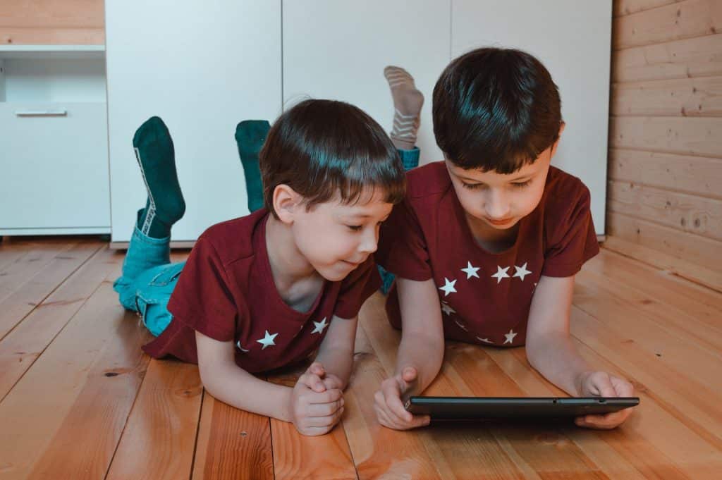 Imagem de dois irmãos gêmeos estudando em casa de forma remota devido à pandemia do Covid-19. Eles estão deitados sobre o chão do quarto. Um deles está segurando o ipad em suas mãos.