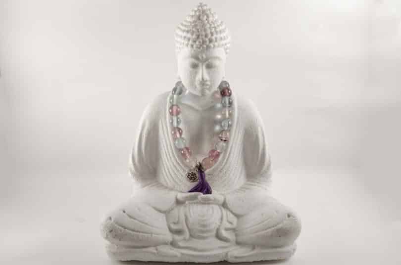 Imagem da estátua de Buddha em posição de meditação. No pescoço dele, uma japamala colorida feita de miçangas.