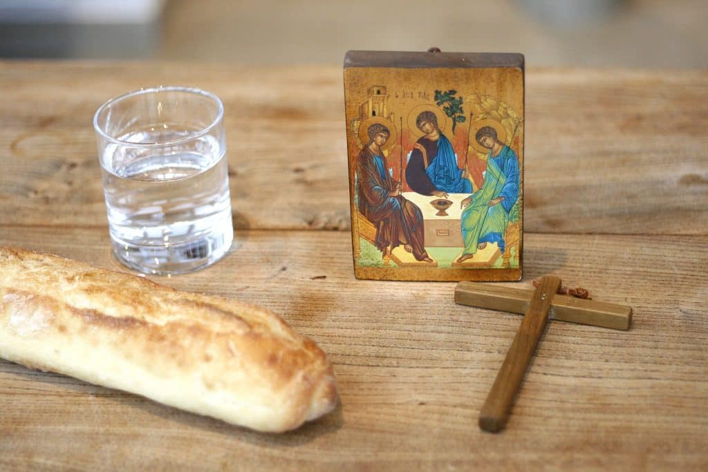 Imagem de uma mesa de madeira. Sobre ela um filão de pão, um copo com água, um crucifixo e um quadrinho pintado com a imagem de Jesus e dois apóstolos. Trata-se de um jejum religioso no período da quaresma.