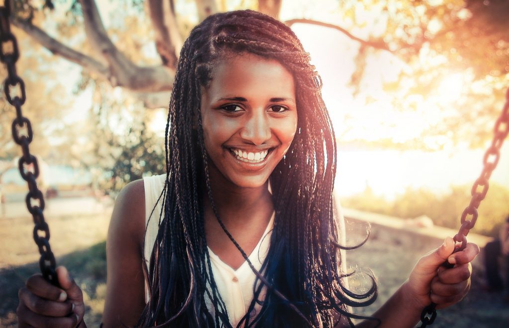 Imagem de uma linda garota negra de cabelos longos sentada em um balanço, apreciando o que há de melhor no verão.
