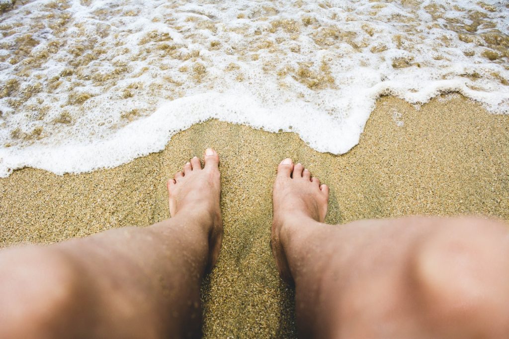 Pés brancos masculinos na areia da praia e onda à frente.