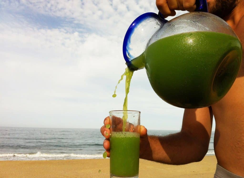 Imagem de um homem na praia segurando um jarro de suco detox verde e em uma das mãos ele segura um copo onde o suco está sendo colocado.