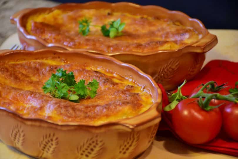 Imagem de duas travessas com lasanhas vegetarianas prontas para serem servidas. Ao lado das travessas dois tomates bem vermelhinhos.