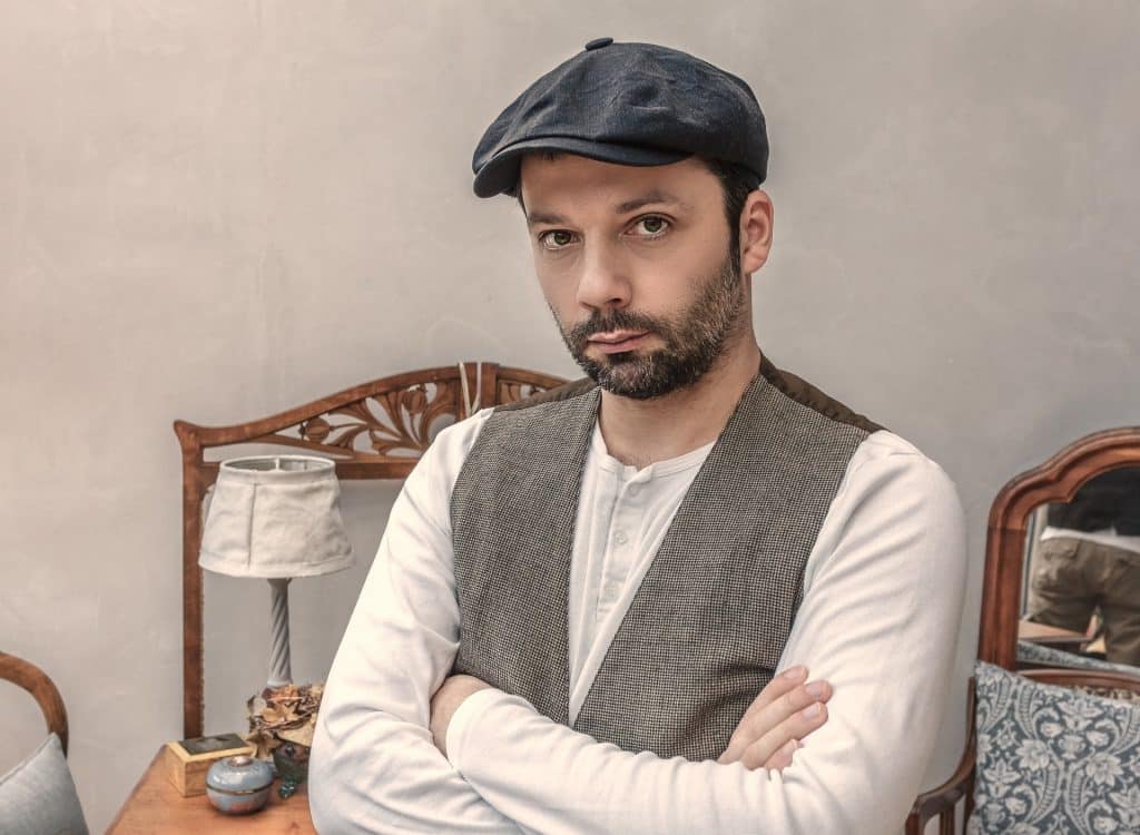 Imagem de um homem de barba cerrada. Ele usa uma camiseta branca de manga longa, uma boina preta e um colete cinza. Ele está de braços cruzados e de mau  humor.