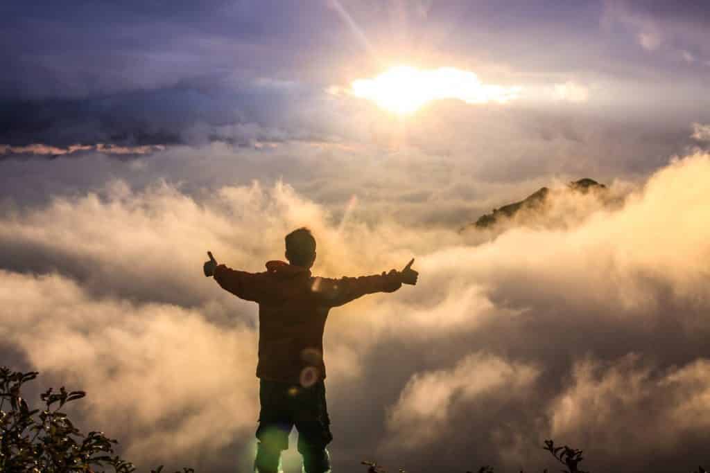 Homem de costas e braços abertos no topo de uma montanha acima das nuvens.