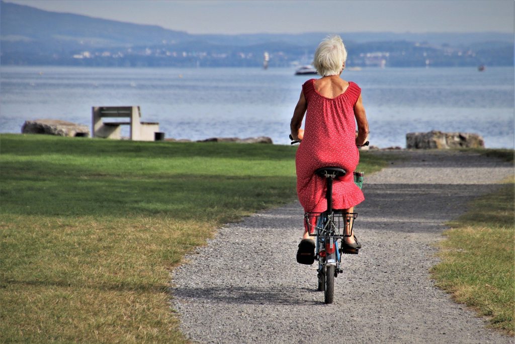 Imagem de uma senhora andando de bicicleta sozinha em uma estrada em direção ao mar. Ela usa um vestido vermelho.