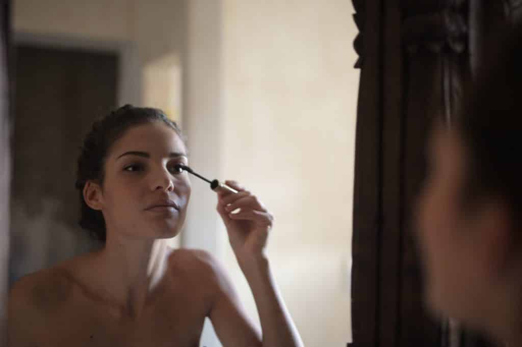Mulher se olhando no espelho e passando maquiagem em um de seus olhos.