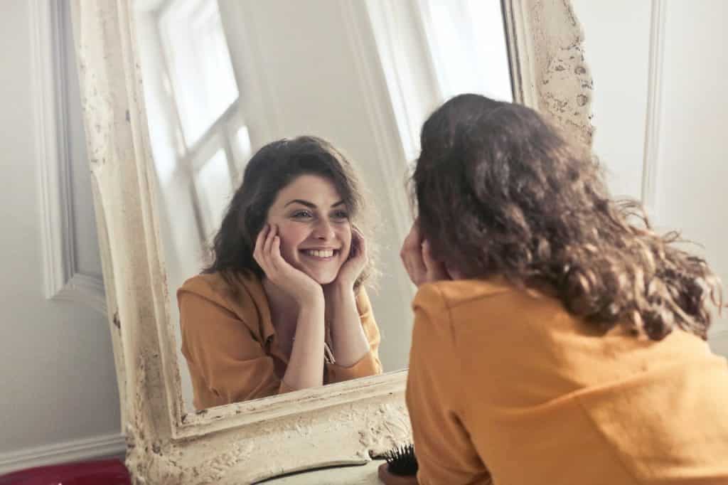 Mulher se olhando no espelho e sorrindo com o rosto apoiado nas mãos, demonstrando felicidade.