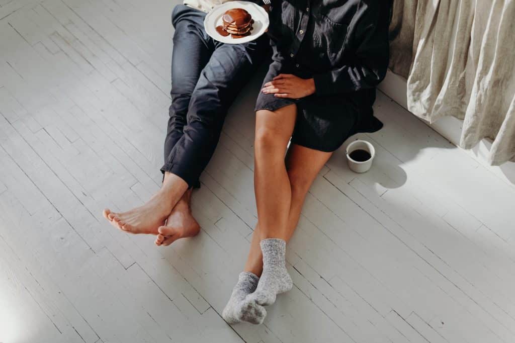 Um homem e uma mulher, mostrados só do tronco pra baixo com um prato de panquecas e chocolate e uma xícara de café - ambos estão sentados no chão.