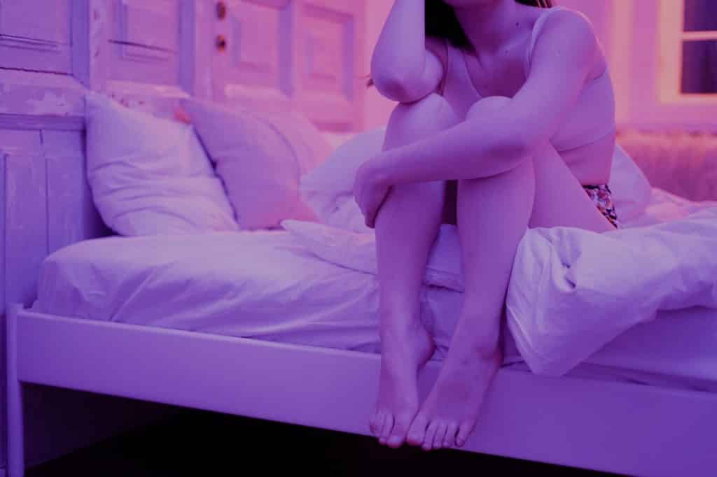 Mulher sentada na cama com o corpo um pouco encolhido, segurando as pernas com um dos braços enquanto o outro cotovelo está apoiado em um de seus joelhos.