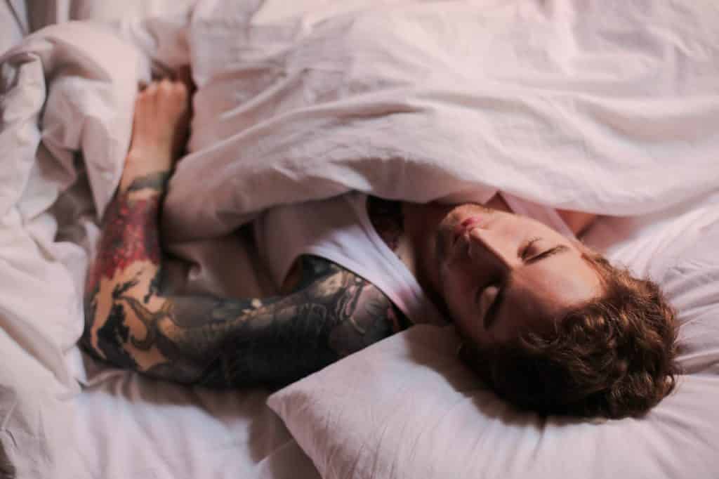 Homem com o braço tatuado deitado e dormindo em uma cama com lençóis brancos.