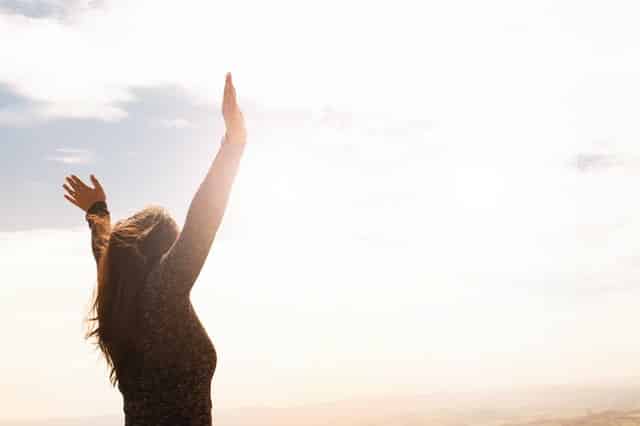 Mulher em pé com braços abertos para o alto e sol refletindo ao fundo