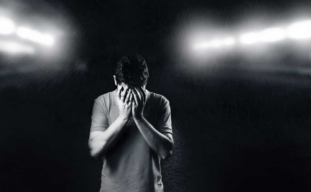 Homem com as duas mãos no rosto remetendo tristeza em meio a um cenário escuro com luzes brancas e distantes.