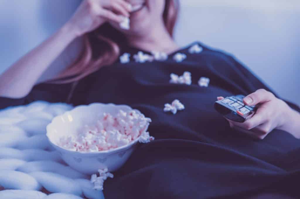 Imagem desfocada de uma mulher comendo pipoca, sorrindo e com um controle de televisão na mão.
