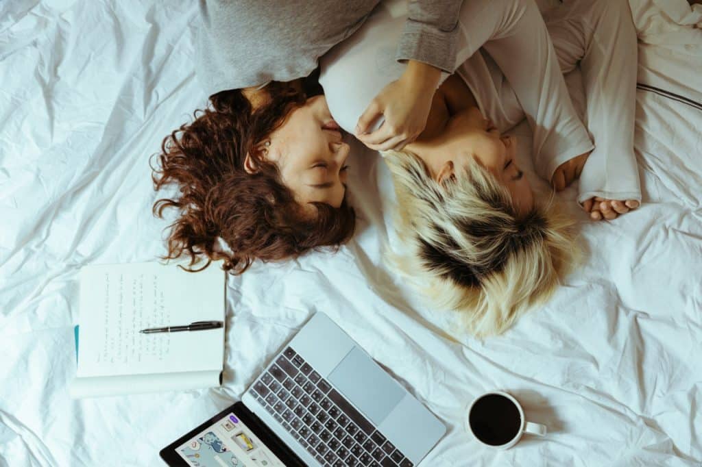 Duas mulheres de cabelos curtos deitadas numa cama branca, com notebook, xícara e caderno ao lado.