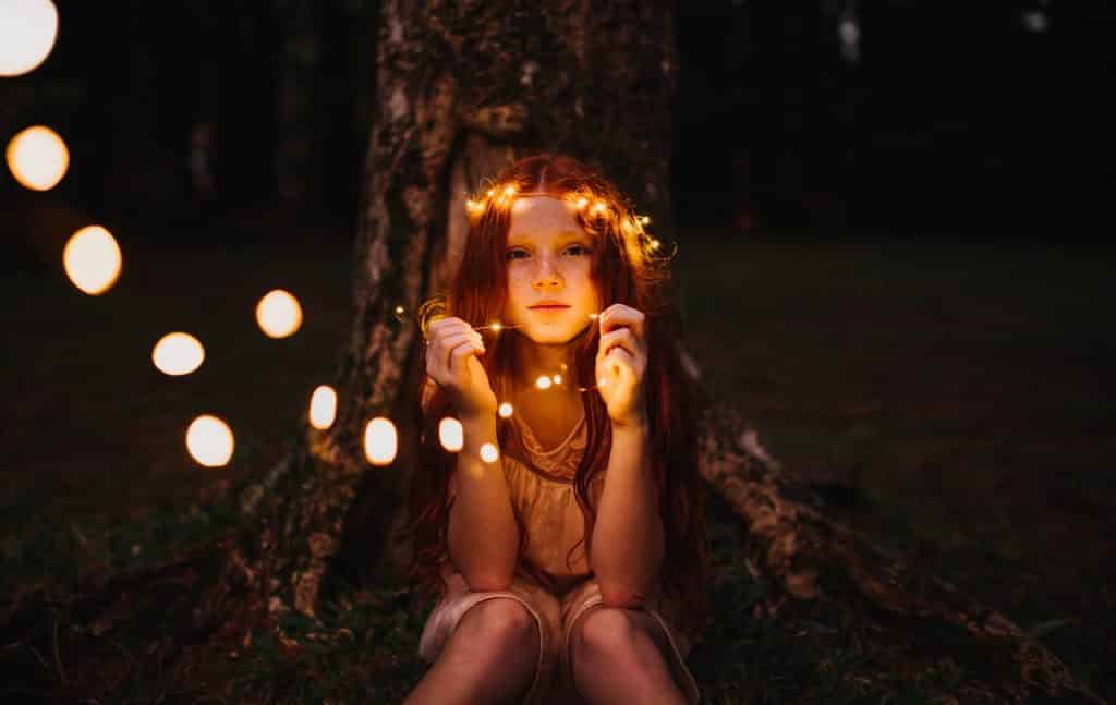 Menina ruiva sentada na base de uma árvore. Ela está segurando luzes pisca-pisca.