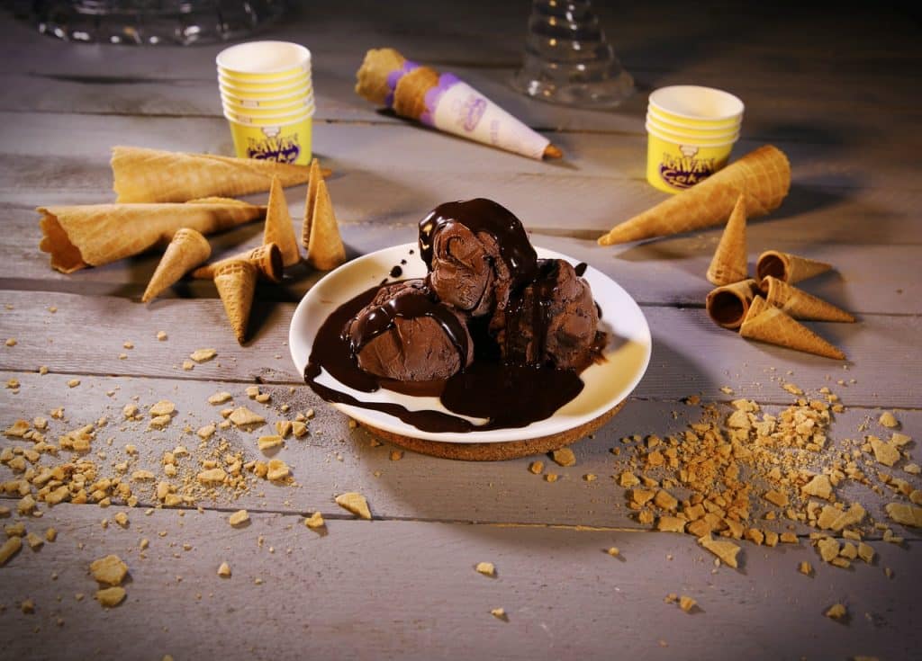 Três bolas de sorvete de chocolate com cobertura ao lado de cones de sorvete espalhados pela mesa.