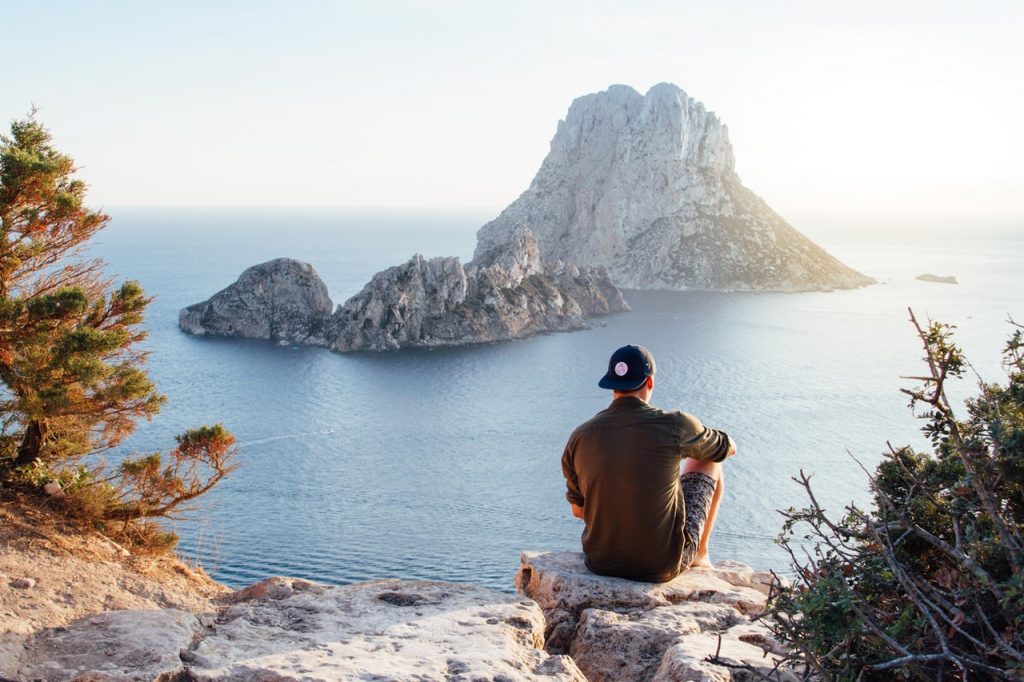 Homem sentado de costas numa pedra, usando boné preto para trás e camiseta verde, observando a praia.