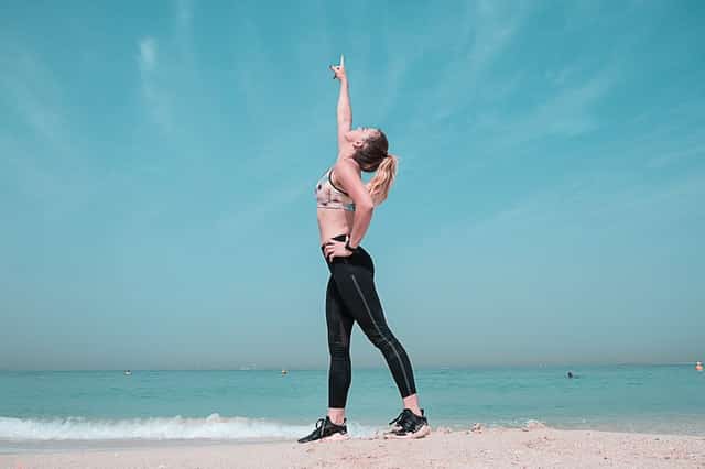 Mulher com roupas de ginástica na praia, com o braço para cima.