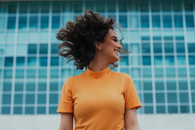 Mulher sorrindo com cabelos ao vento e janelas de prédio ao fundo