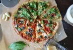 Imagem de uma linda e saborosa pizza vegetariana já fatiada. Ao lado dela alguns ingredientes como: manjericão, alho e queijo.