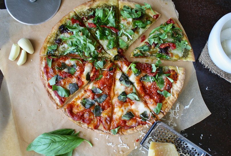 Imagem de uma linda e saborosa pizza vegetariana já fatiada. Ao lado dela alguns ingredientes como: manjericão, alho e queijo.