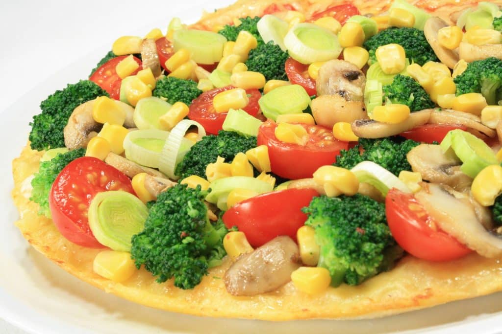 Imagem de uma pizza de brócolis com milho e outros ingredientes como tomate e alho poró.