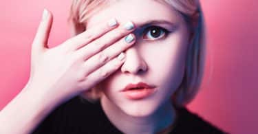 Imagem do rosto de uma jovem. Ela tampa um dos seus olhos. Suas unhas estão pintadas de cores diferentes de esmate.