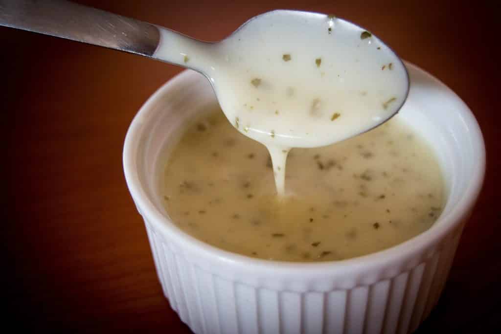 Ramekin com molho branco dentro e uma colher com um pouco de molho escorrendo.