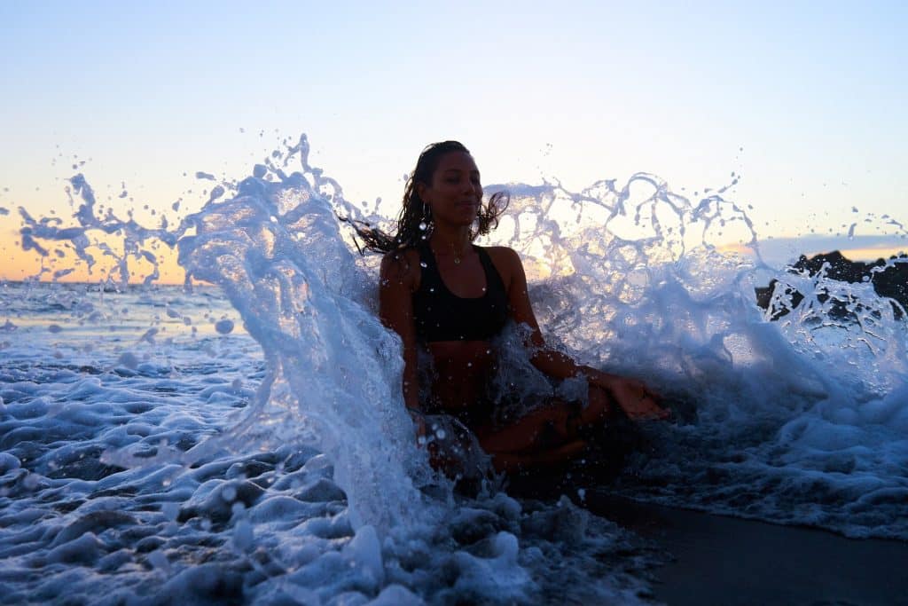 Imagem de uma mulher sentada na areia, bem próximo à beira do mar. Ela está meditando e as ondas do mar estão quebrando em suas costas.
