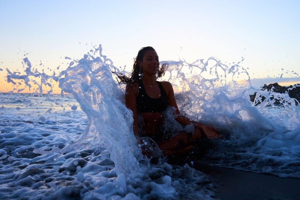 Imagem de uma mulher sentada na areia, bem próximo à beira do mar. Ela está meditando e as ondas do mar estão quebrando em suas costas.
