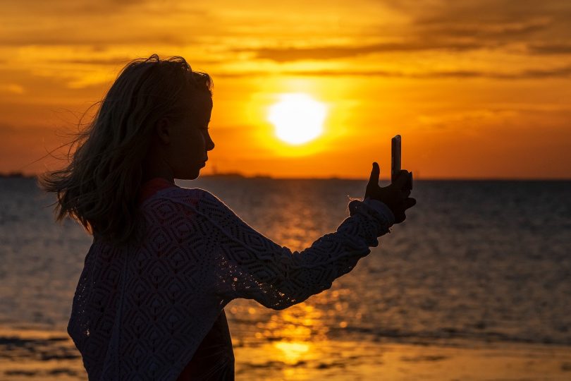 Imagem da silhueta de uma jovem fazendo uma selfie em pleno por do sol em um alto dia de verão.