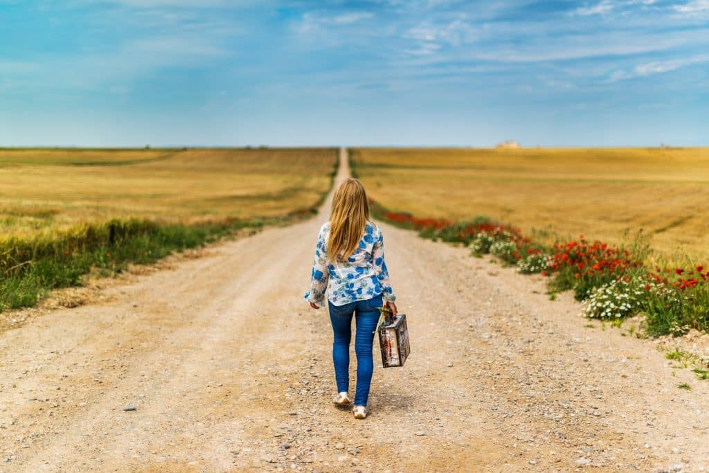 Imagem de uma extensa estrada de terra cercada por um campo verde. Nela, caminha uma mulher sozinha segurando em uma das mãos uma mala.