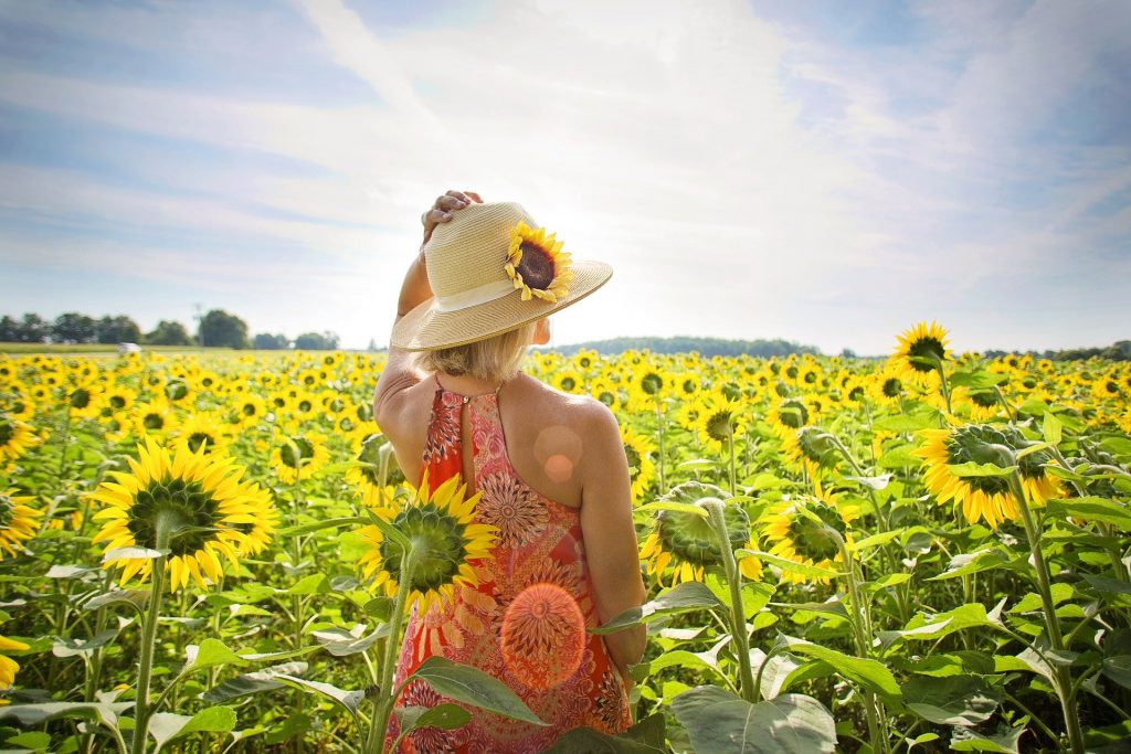 Imagem de um campo de girassóis e nele está uma mulher segurando um chapéu de palha sobre a sua cabeça. Ela está curtindo o verão aceitando a sua melhor forma.