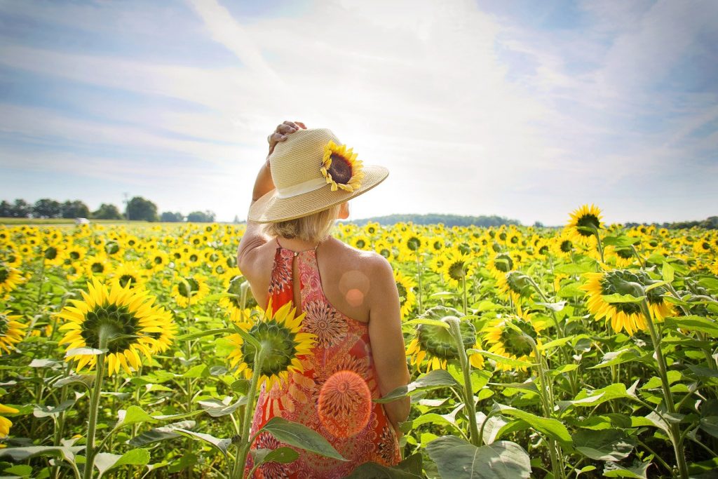 Imagem de um campo de girassóis e nele está uma mulher segurando um chapéu de palha sobre a sua cabeça. Ela está curtindo o verão aceitando a sua melhor forma.
