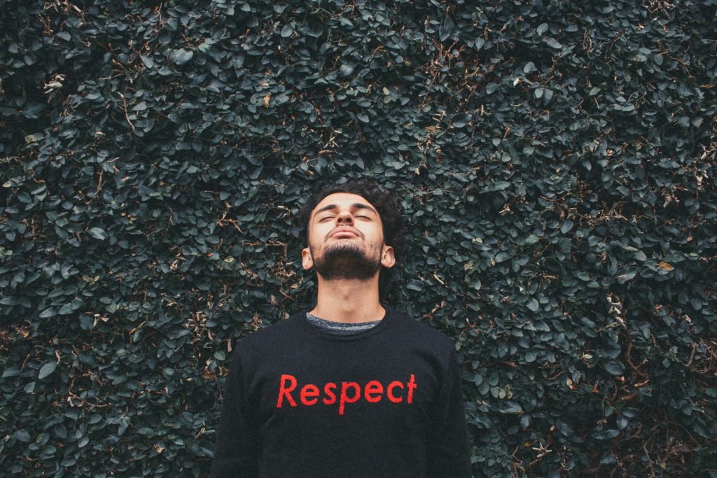 Homem branco de barba preta, cabeça tombada para trás, usando moletom com a palavra "respect" em vermelho, em frente à uma parede de folhas.